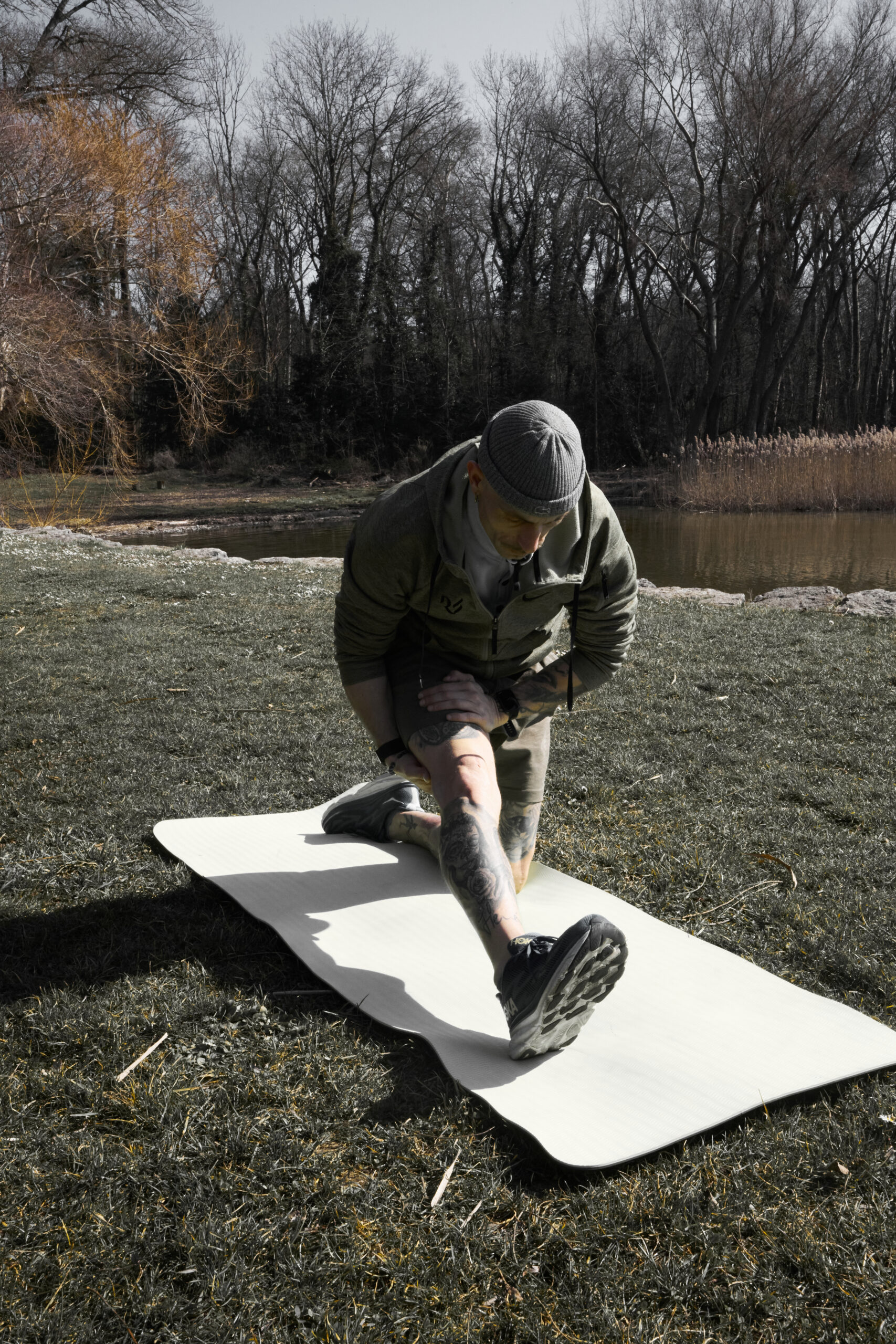 Coach faisant un étirement en plein air sur un tapis de sol, près d’un lac, vêtue d’une tenue de sport et d’un bonnet, concentrée sur un exercice de mobilité.